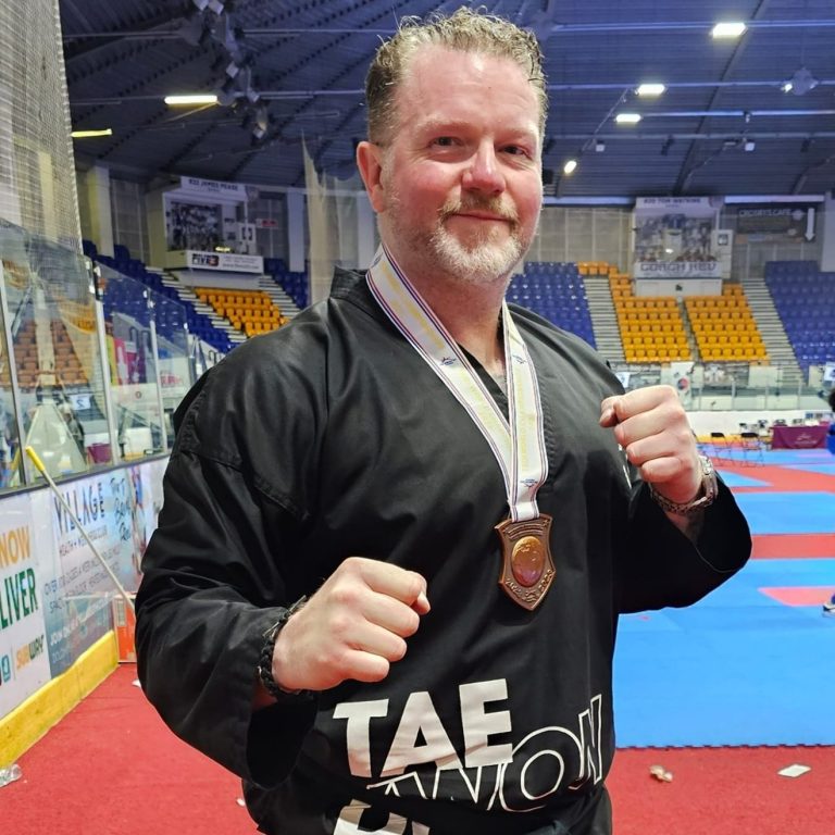 Kevin Wyllie Kevin Whyllie wearing a black uniform and medal, posing confidently in a sports hall.